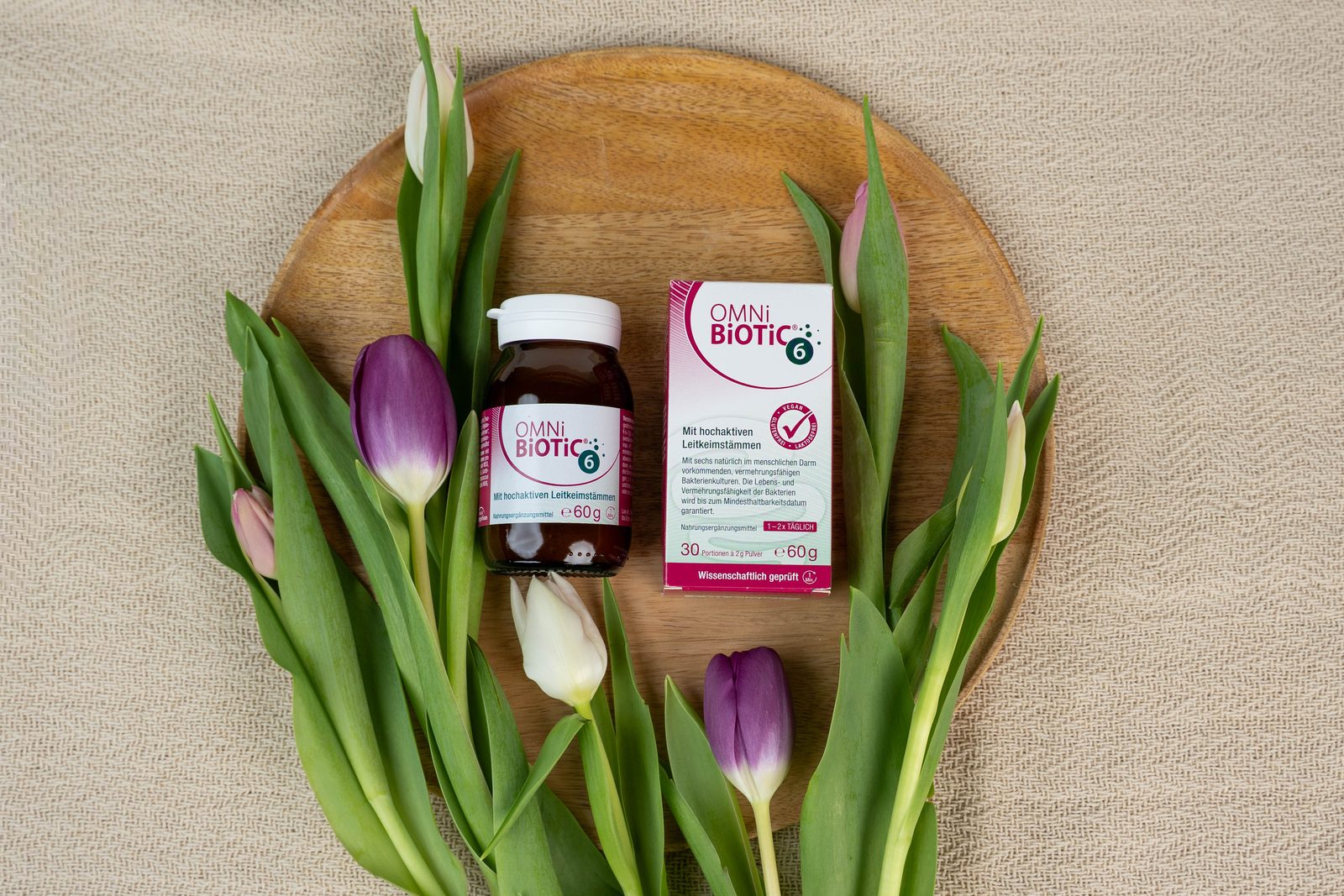 OMNi-BiOTiC 6 Dose und Packung auf einem Holzteller, daneben Tulpen